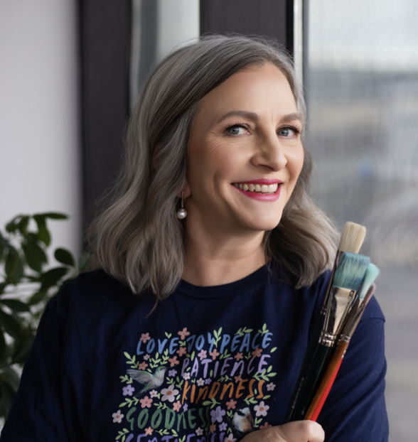 A smiling woman with gray hair holding paintbrushes, standing near a window with a blurred outdoor view.