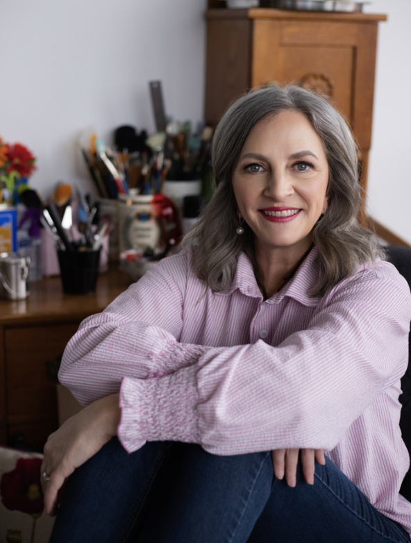 A smiling woman with gray hair, wearing a pink striped shirt, sitting in a home office or craft room with various art supplies and a wooden cabinet in the background.