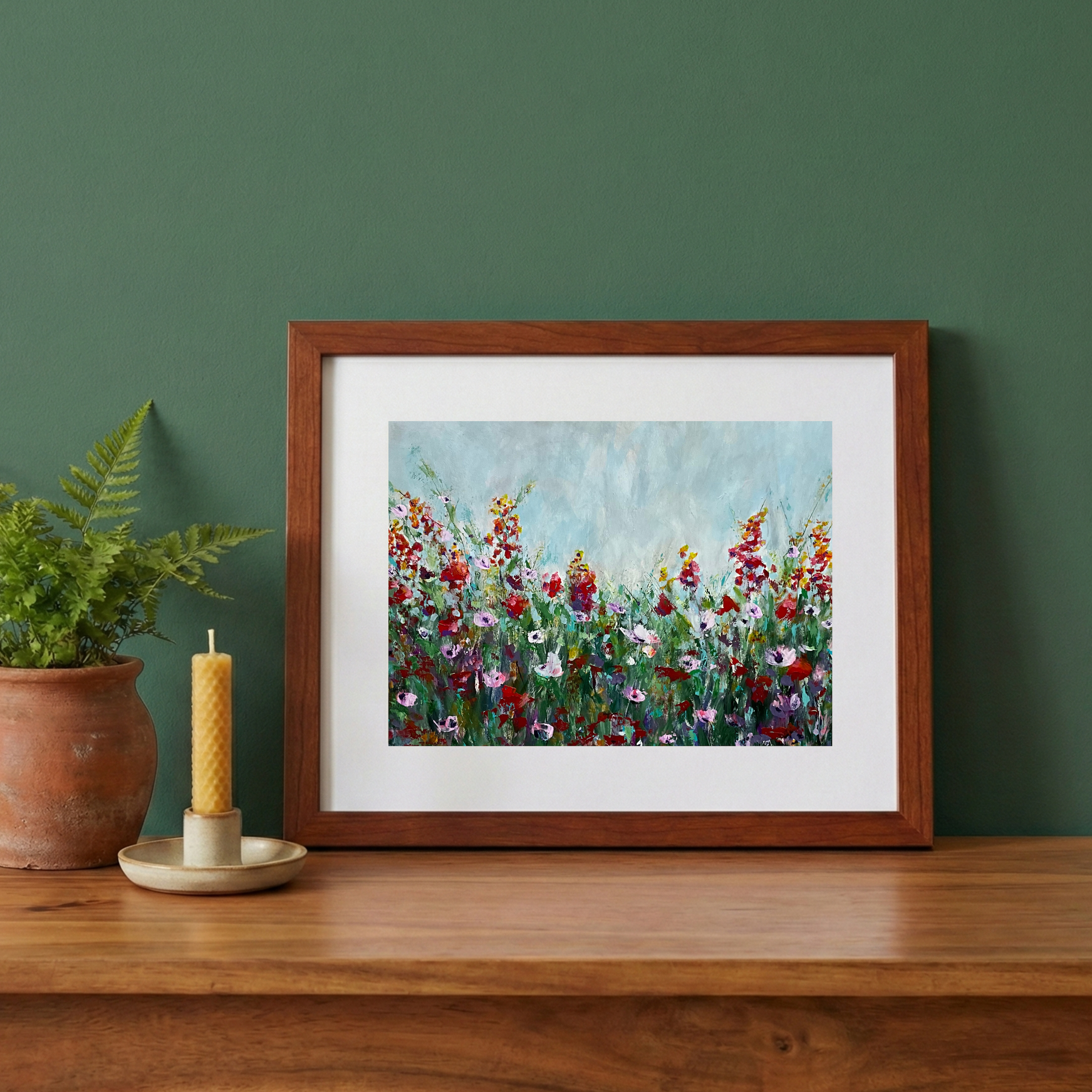 Framed floral painting on a green wall, with potted fern plant and yellow candle on a wooden surface.
