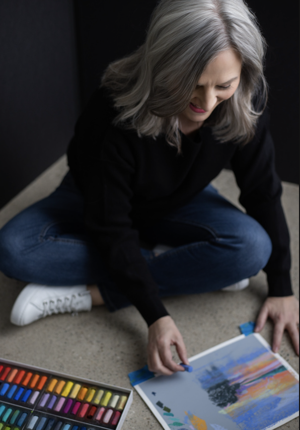 A woman with gray hair sitting cross-legged on the floor, smiling while using a set of colorful pastel chalks and creating a landscape painting with a sunset sky and trees.