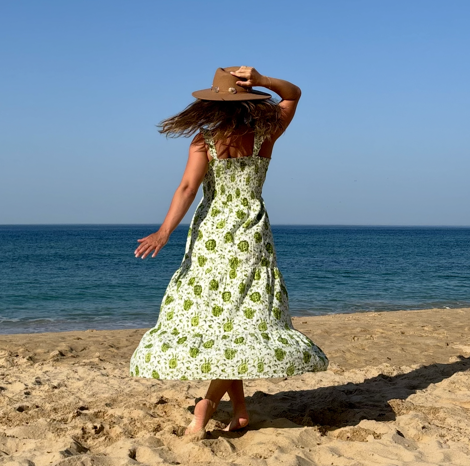 Vrouw in zomerjurk en hoed die op het strand staat, met de zee op de achtergrond.