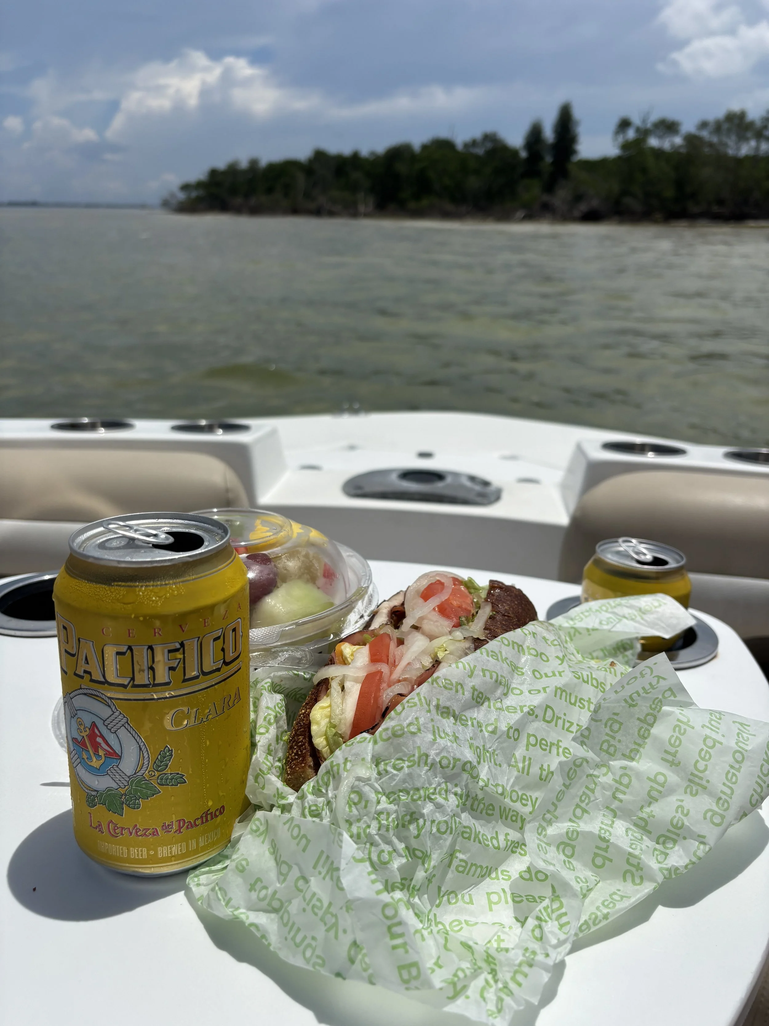 A boat deck with two cans of Pacifico beer, a sandwich with tomatoes, lettuce, and meat wrapped in green and white paper, and a bowl of grapes, with a body of water and a tree-lined shoreline in the background.