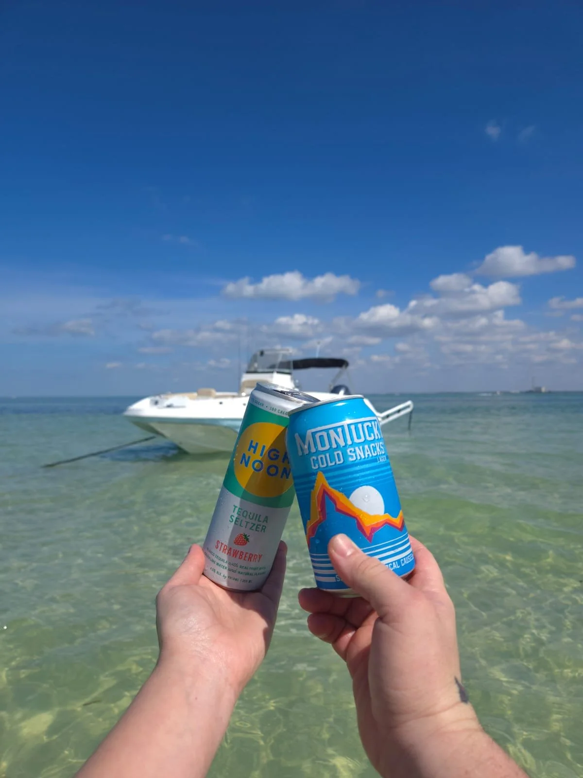 Two hands holding cans of drinks over clear ocean water with a white boat in the background and a blue sky with scattered clouds.