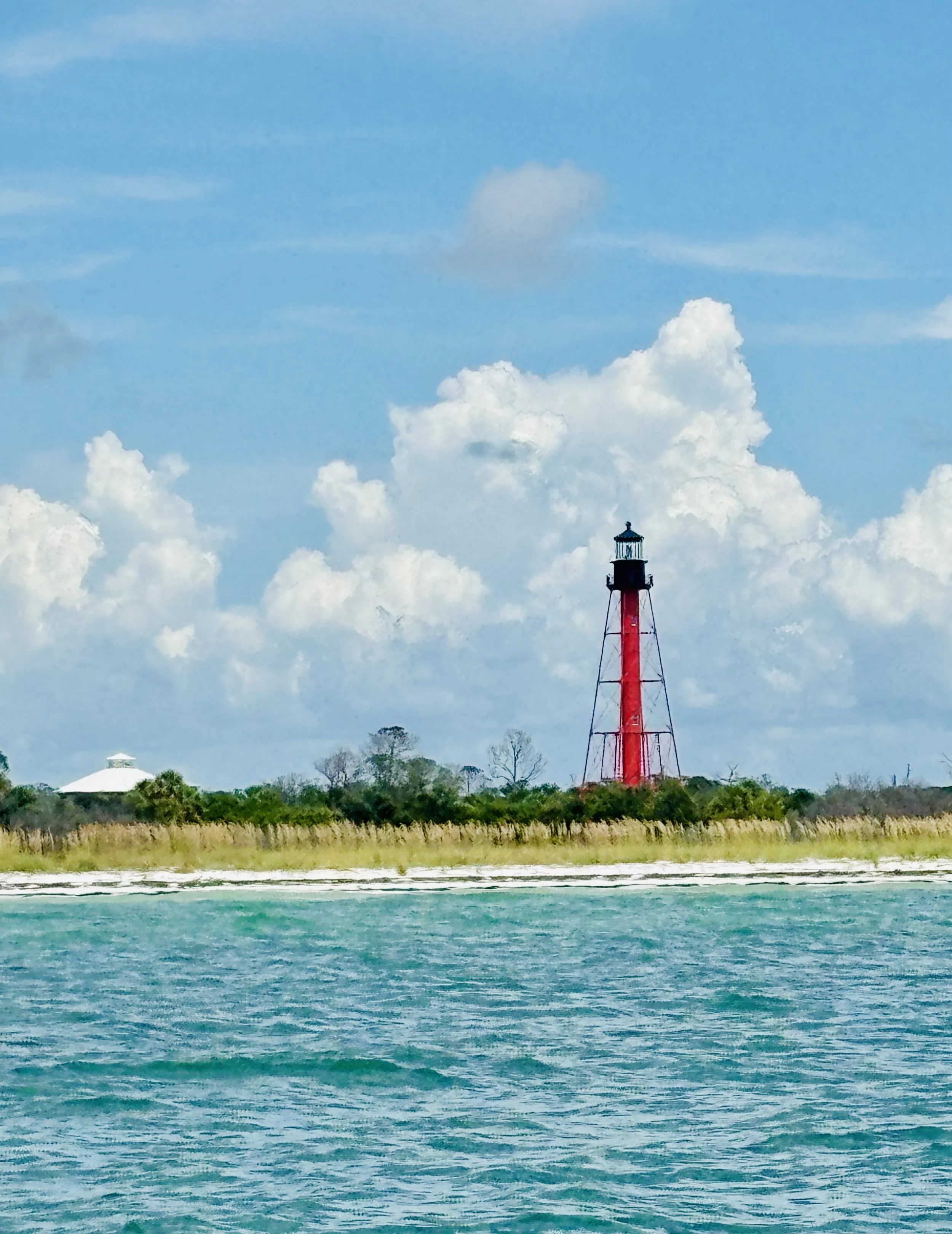 A red lighthouse on a grassy shoreline with a blue sky and white clouds, water in the foreground.