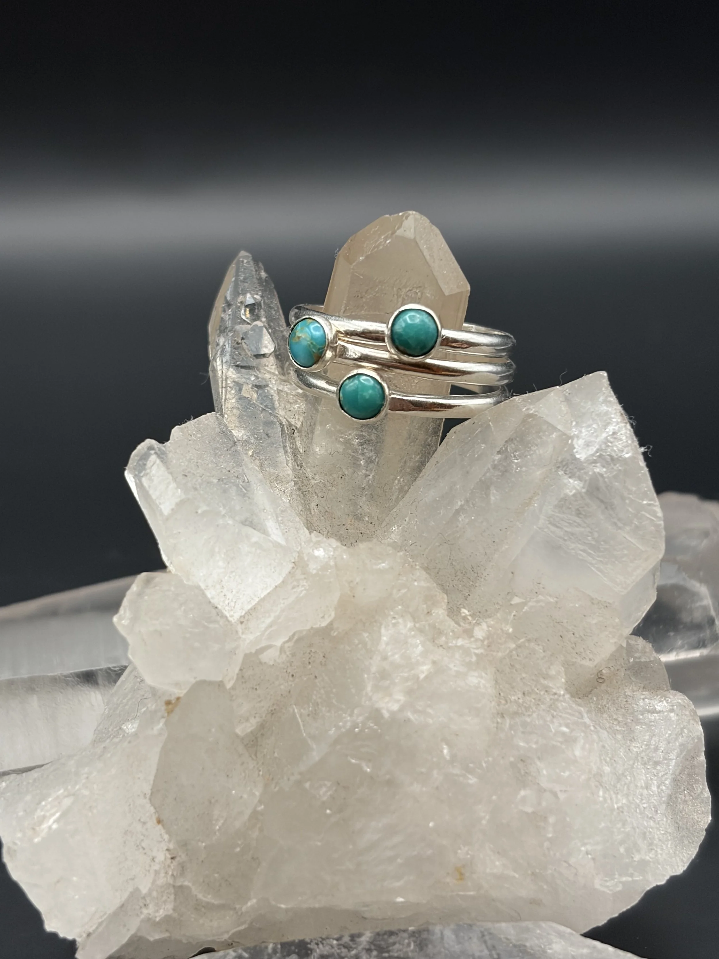 A silver ring with three turquoise stones, resting on a cluster of translucent, irregularly shaped crystals.