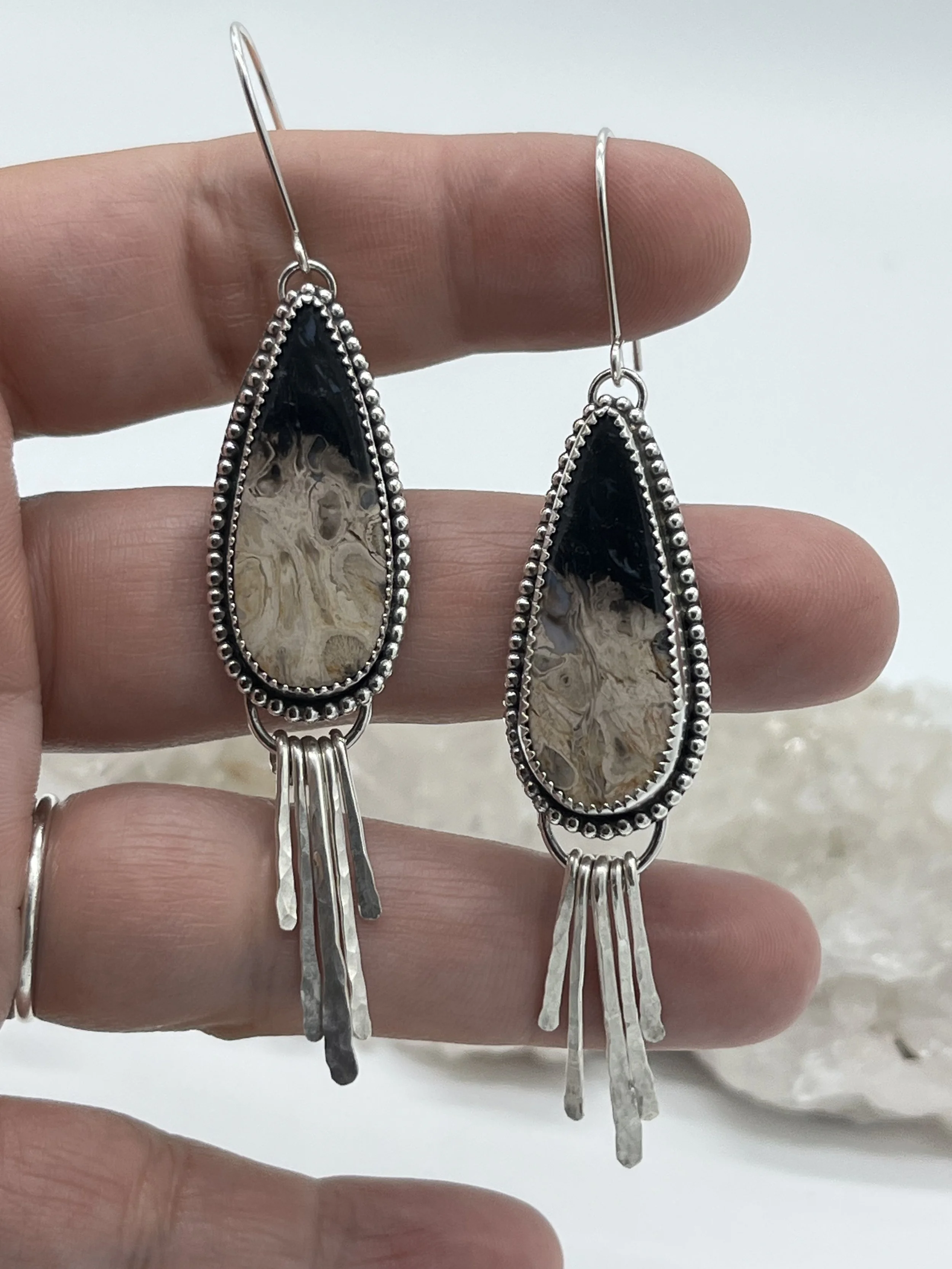Pair of silver dangle earrings with teardrop-shaped black and beige patterned stones, decorated with a beaded border and hanging silver strands.