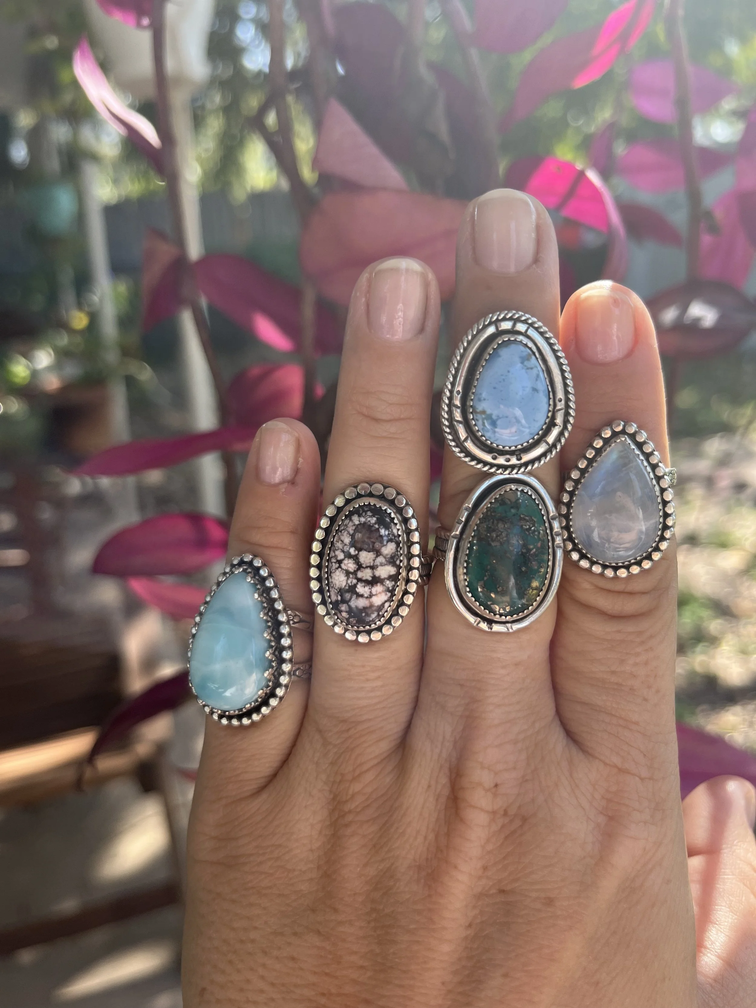 A hand wearing five large silver rings with colorful stones, set against a background of pink and green leaves.