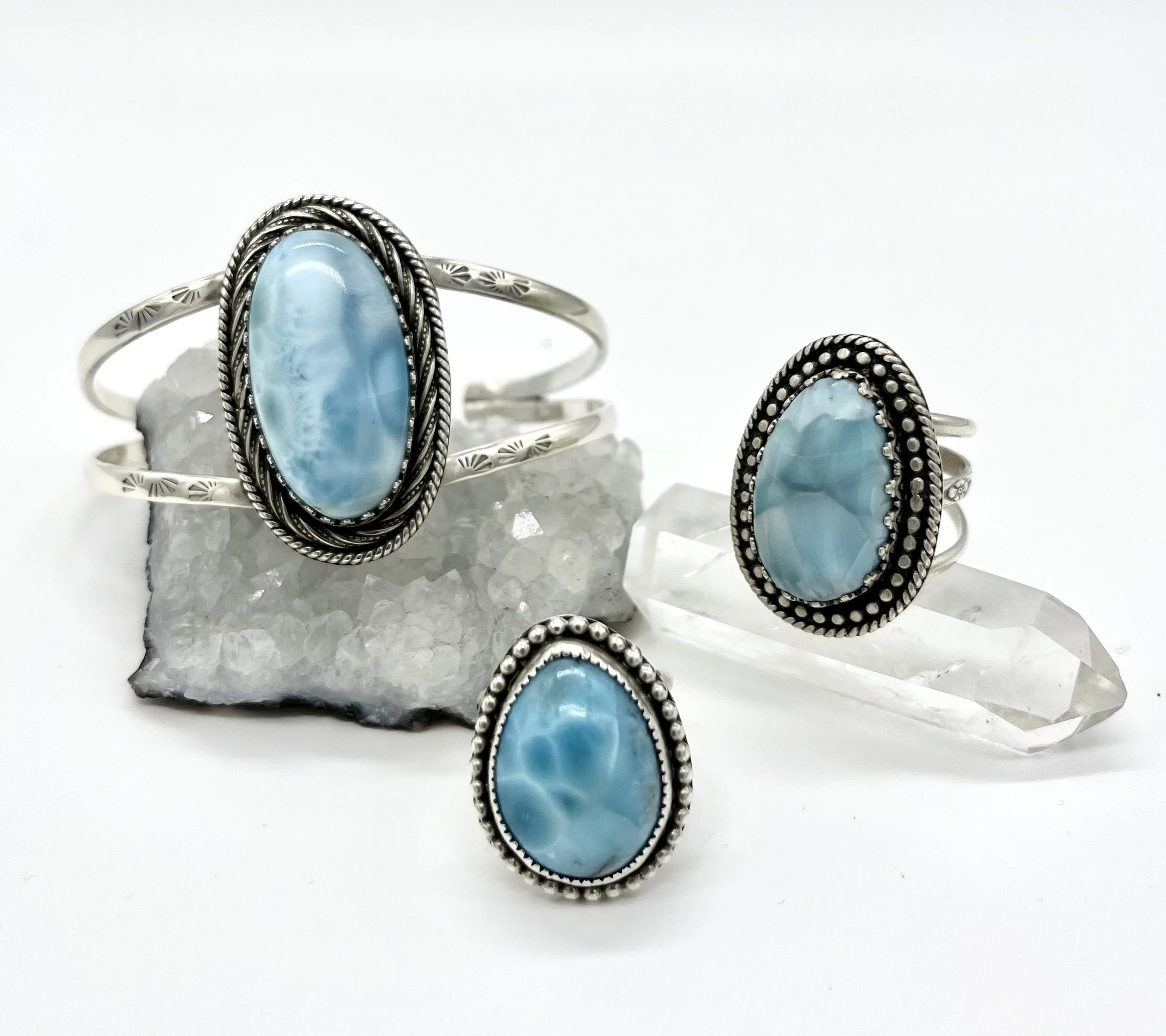 Silver jewelry set with large oval blue gemstones, including a ring, bracelet, and ring, displayed on rocks and a clear quartz point.