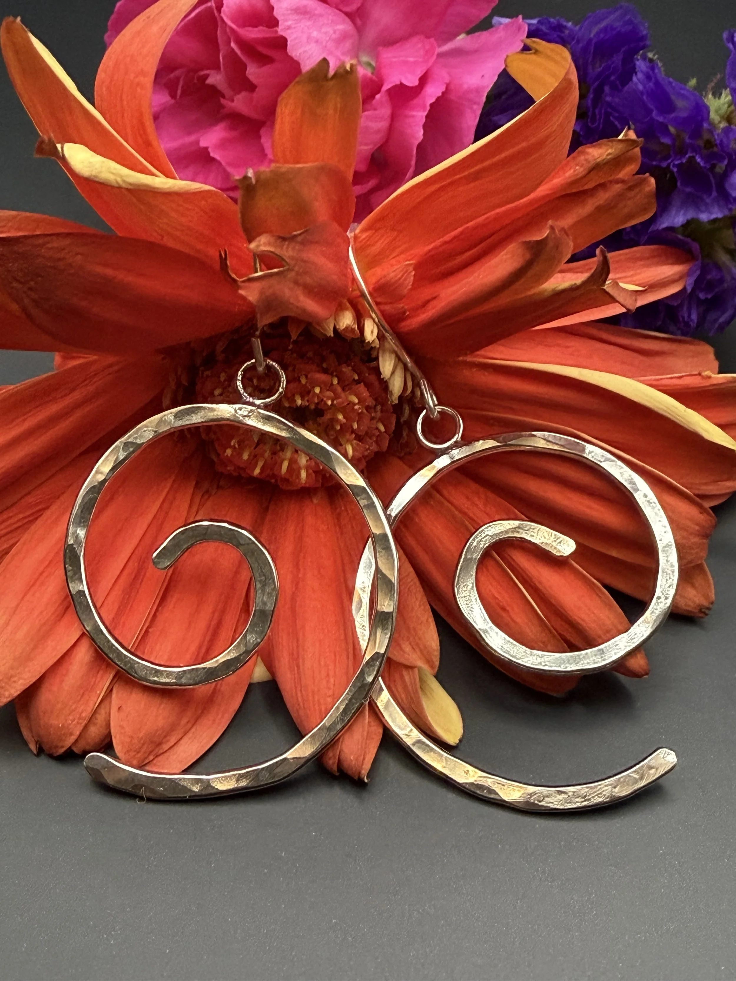 Silver earrings shaped as a word with swirling curves, placed on orange and pink flowers with purple accents.