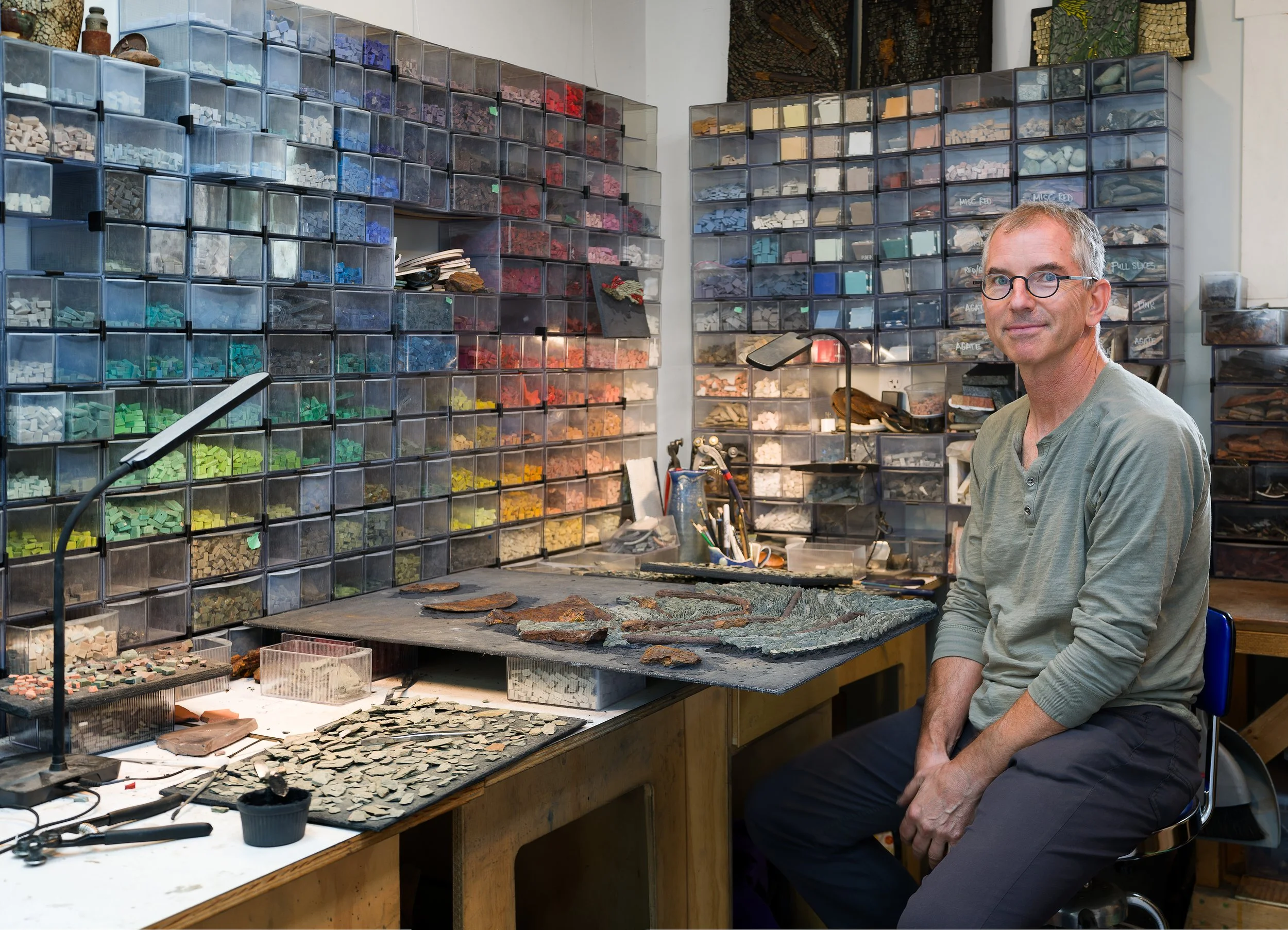 A man sitting next to a workbench covered with archaeological artifacts, in front of storage bins filled with colored stones and materials.