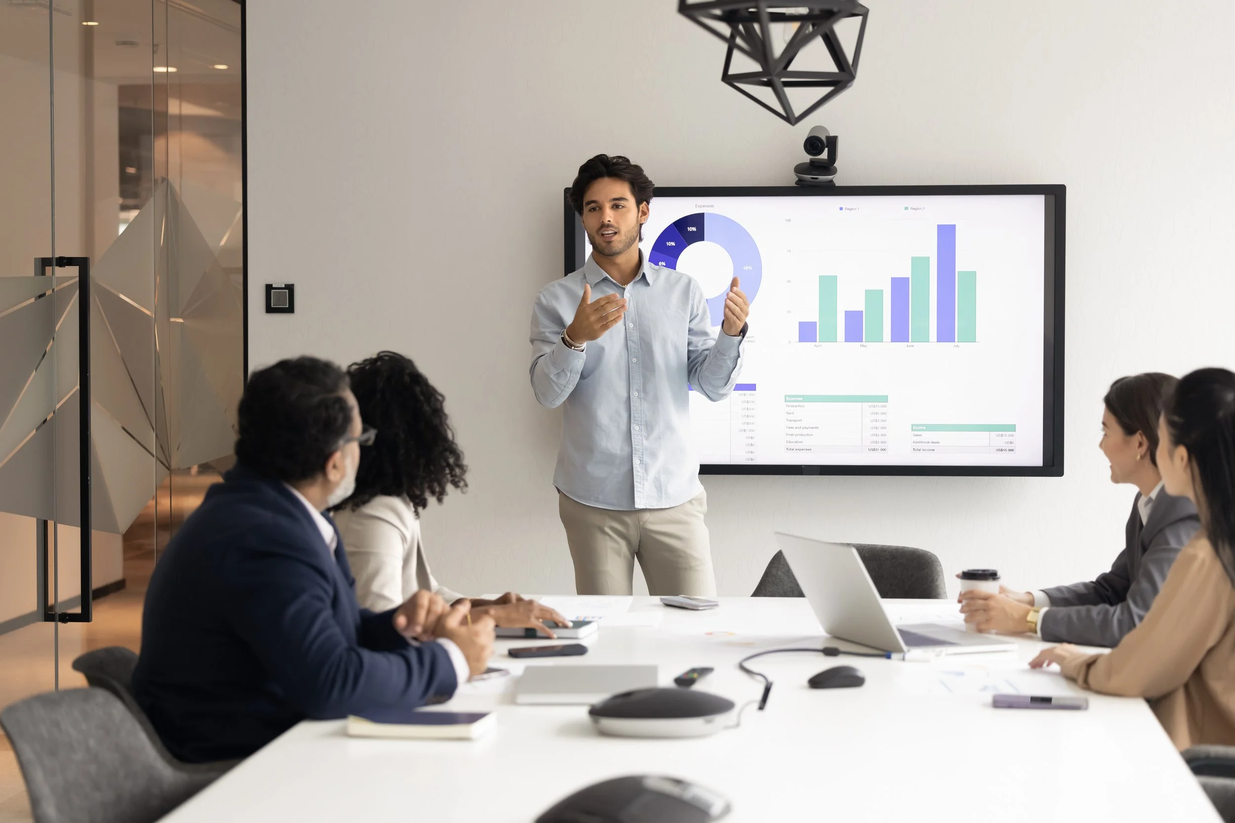 A man giving a presentation in a conference room with five people seated around the table. The man is gesturing with his hands in front of a large screen displaying charts and graphs. The seated people are listening and have laptops and notebooks in front of them.