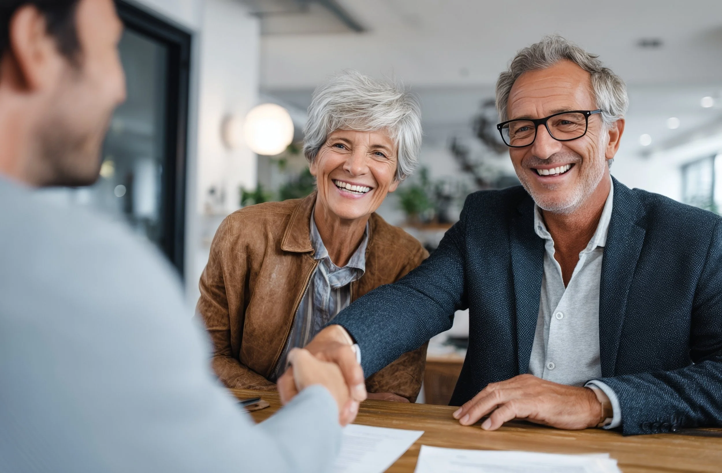 A happy senior couple shaking hands with a man across a table in a modern office or meeting space, smiling warmly.