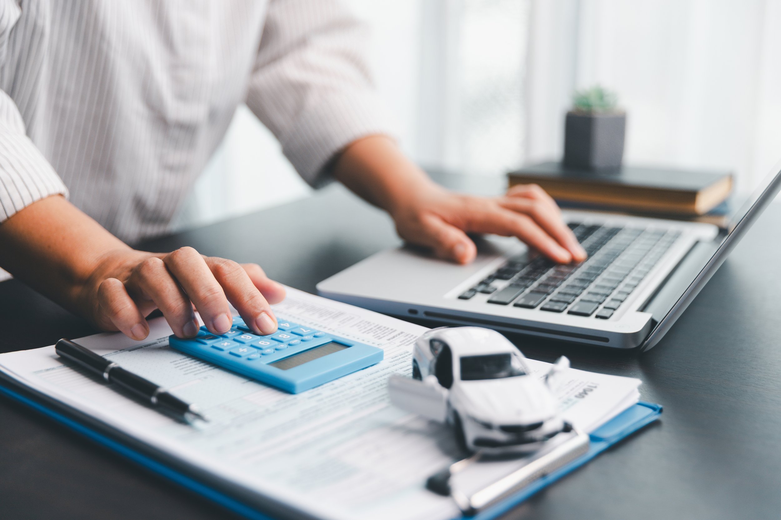 Person working at a desk with a computer, blue calculator, pen, and a car model on top of documents.