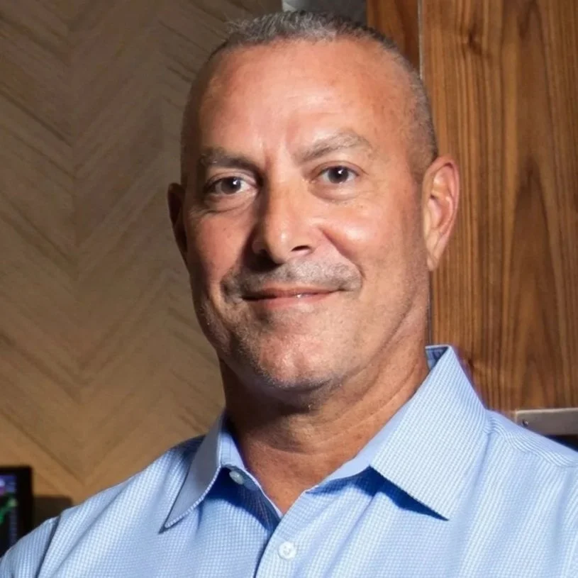 A middle-aged man with short hair and a slight smile, wearing a light blue collared shirt, standing indoors with a wooden wall in the background.