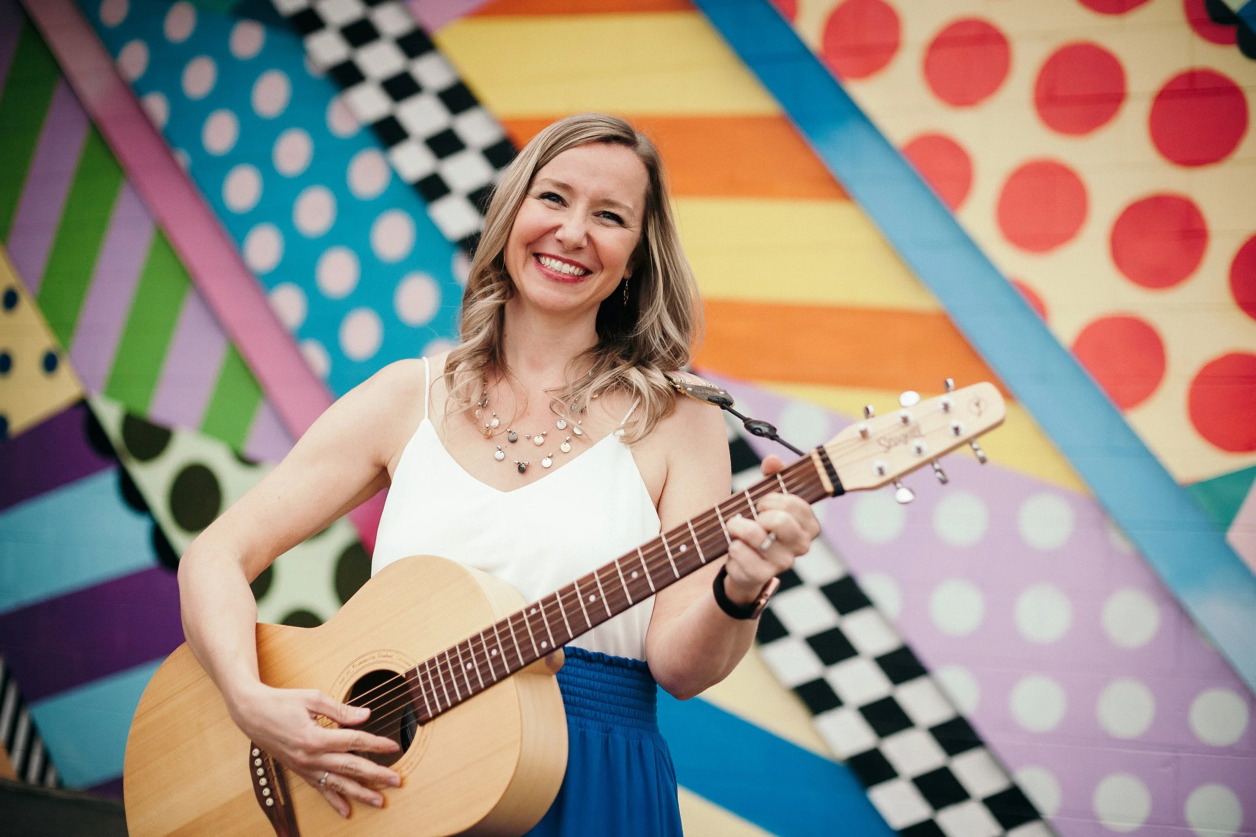 Jaime Lawrence, A woman with blonde hair, wearing a white tank top and a blue skirt, playing an acoustic guitar in front of a colorful, geometric mural (Brightwalls Jackson, MI).