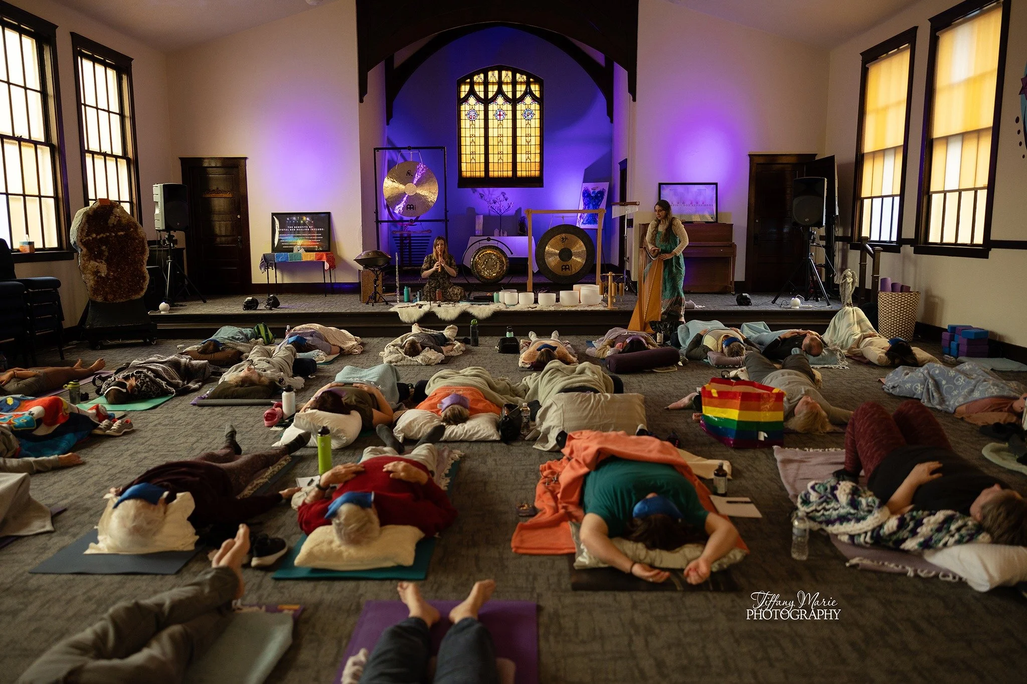 People in a sound healing and meditation class lying on mats and pillows with their eyes closed in a spacious room with large windows, before a stage with musical instruments and a stained glass window.