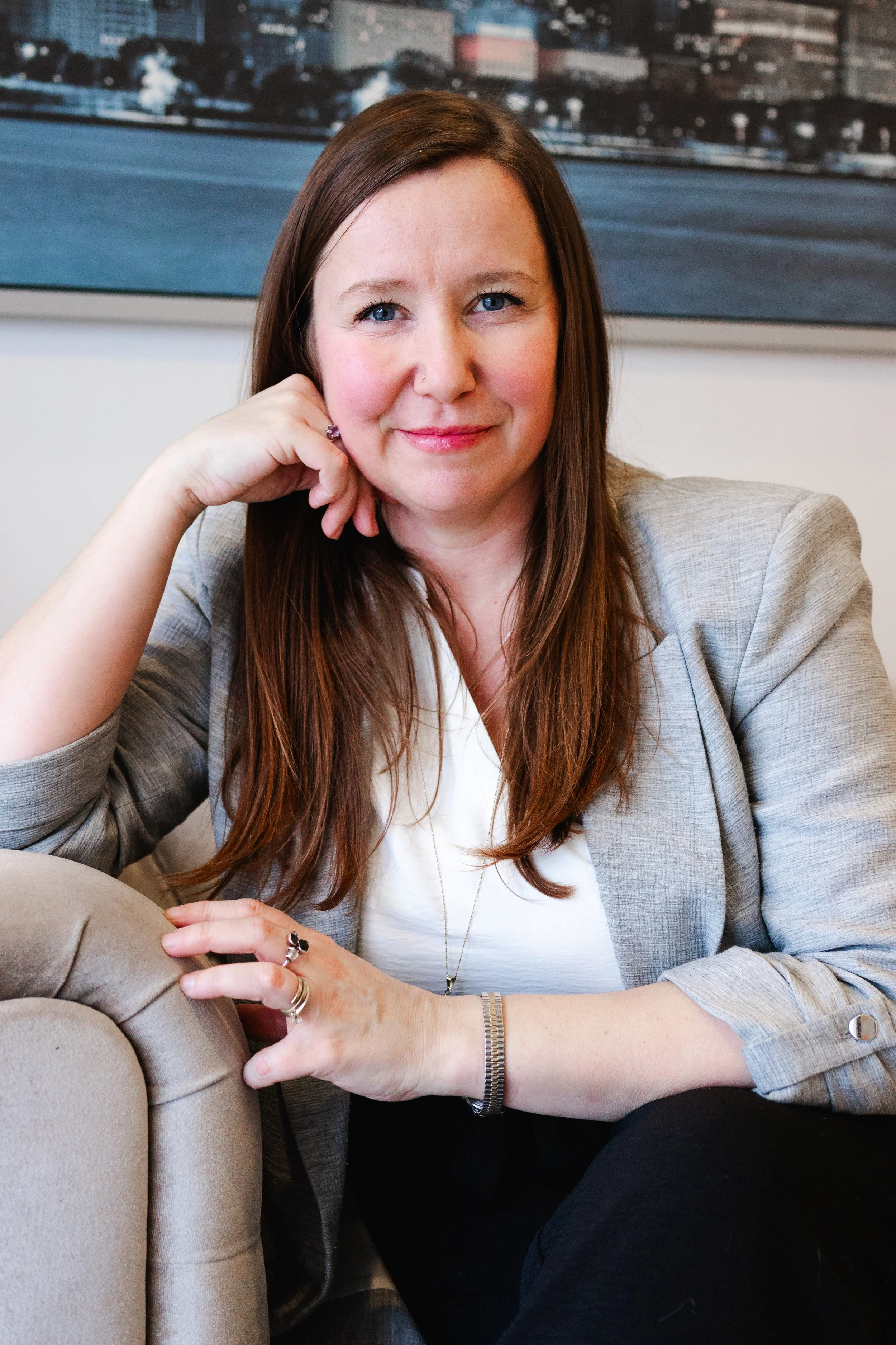 Jaime Lawrence, a woman with long blonde hair, wearing a white top, smiling, sitting on a mustard-colored leather couch with a silver MacBook on her lap, in front of a brick wall and green plants.