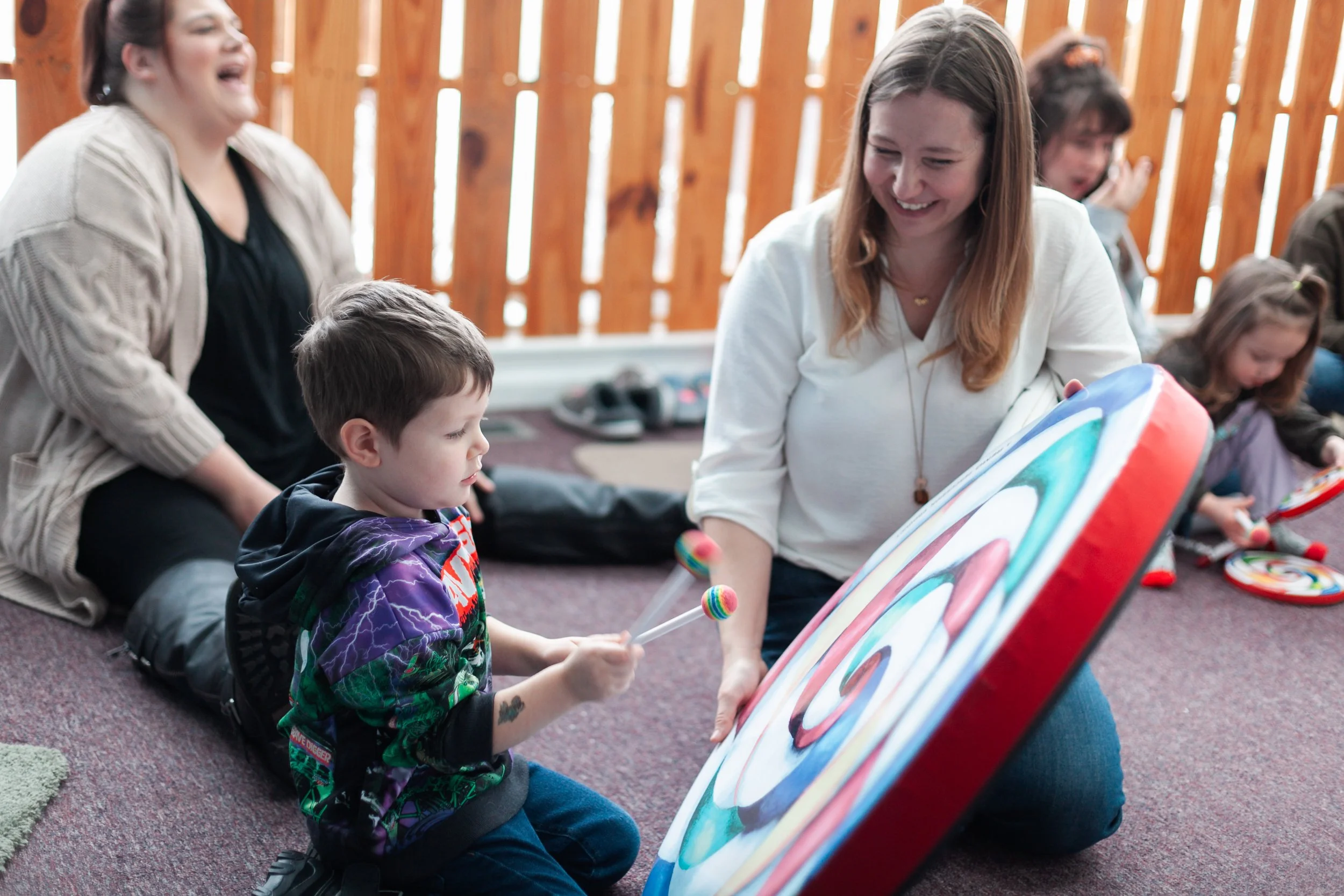 A group of children and adults playing a large colorful lollipop drum inside a room with wooden walls.