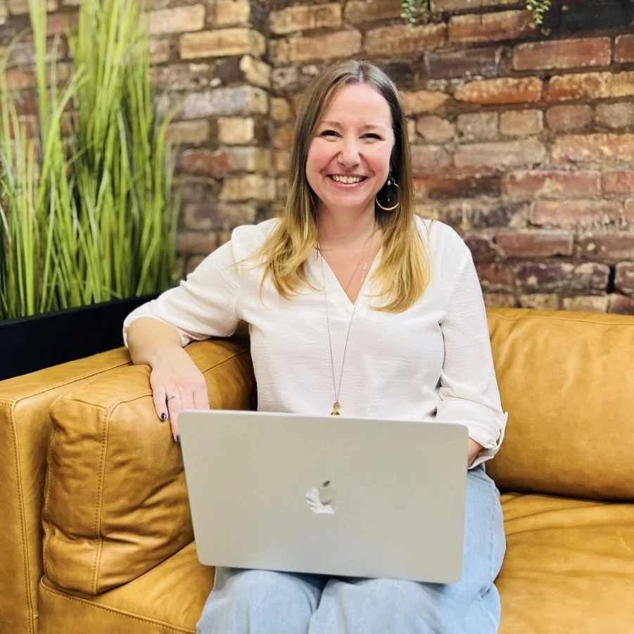 Jaime Lawrence, a woman with long blonde hair, wearing a white top, smiling, sitting on a mustard-colored leather couch with a silver MacBook on her lap, in front of a brick wall and green plants.