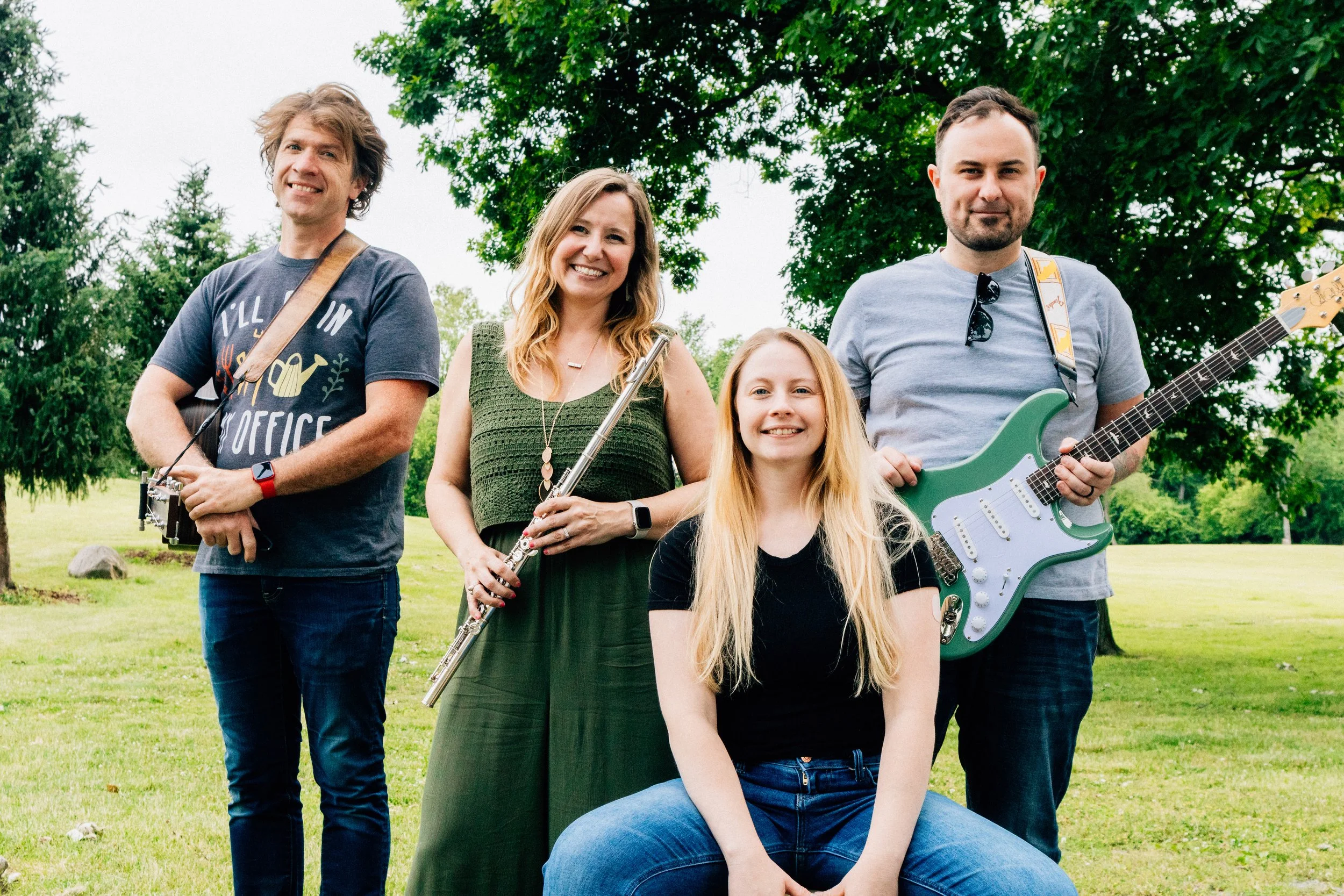 Group of four people outdoors, holding musical instruments, smiling at the camera on a grassy field with trees. The Harmony Gardeners