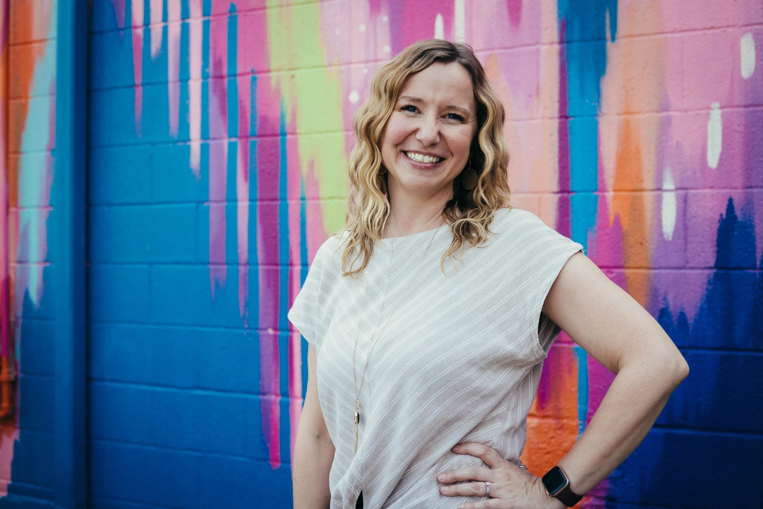 Jaime Lawrence, a woman with wavy blonde hair smiling and standing in front of a colorful graffiti wall (Bright Walls downtown Jackson, MI)