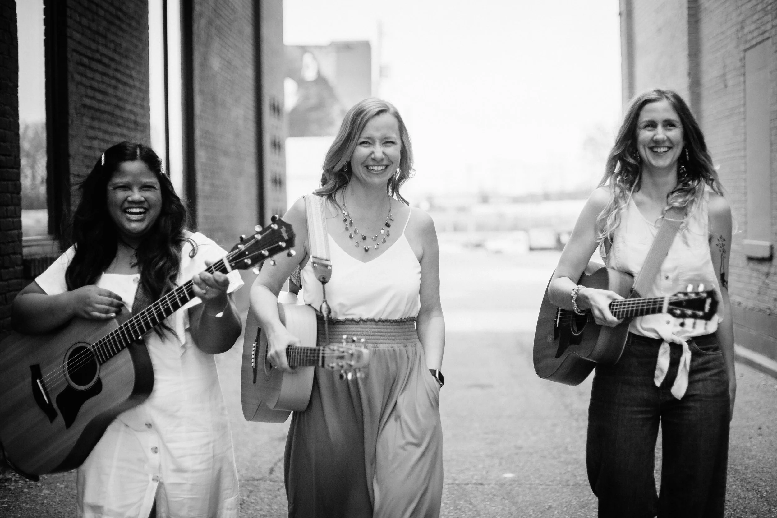Three women walking down a narrow alley, playing guitars and smiling, in a black and white photo.