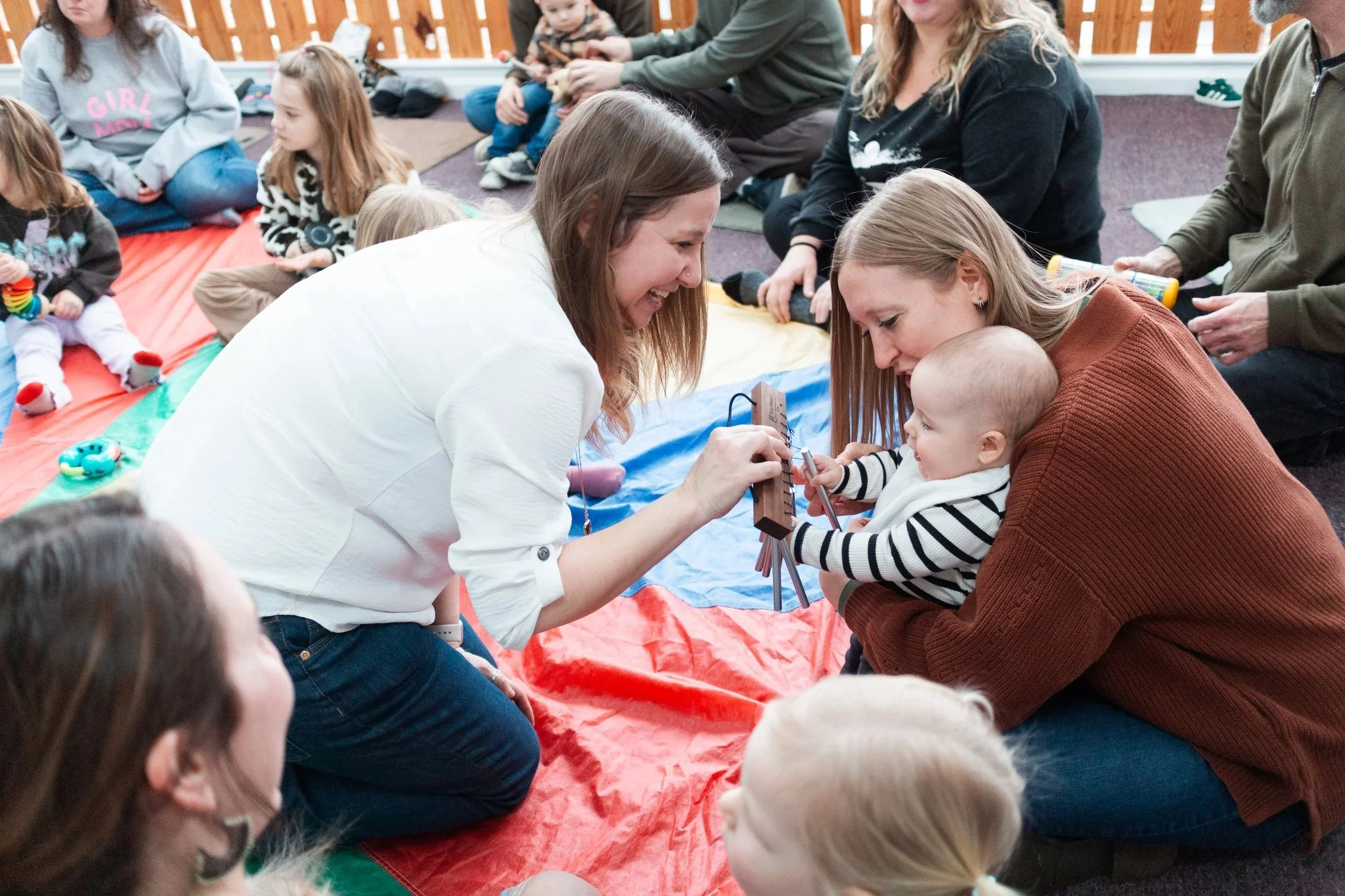 Adults and children participating in an early childhood music group activity indoors, some sitting on blankets, with two women engaging with a baby in the foreground.