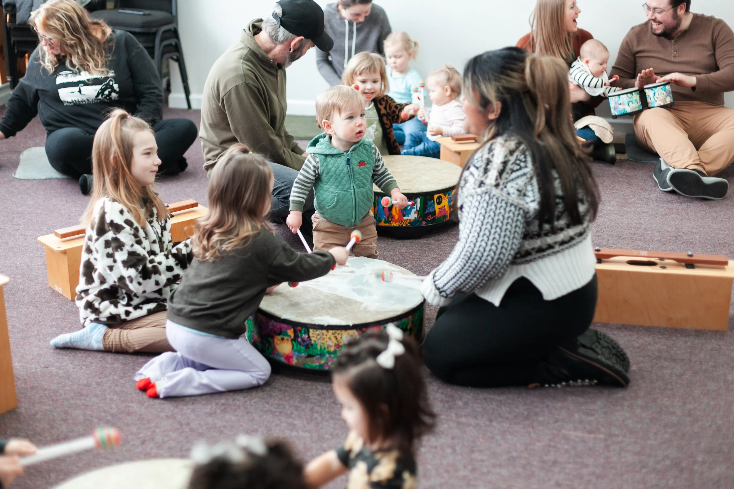 Children and adults in a Harmony Garden Sprouts early childhood music class, sitting on the floor in a circle, playing musical instruments and engaging in a musical activity in a casual indoor setting.