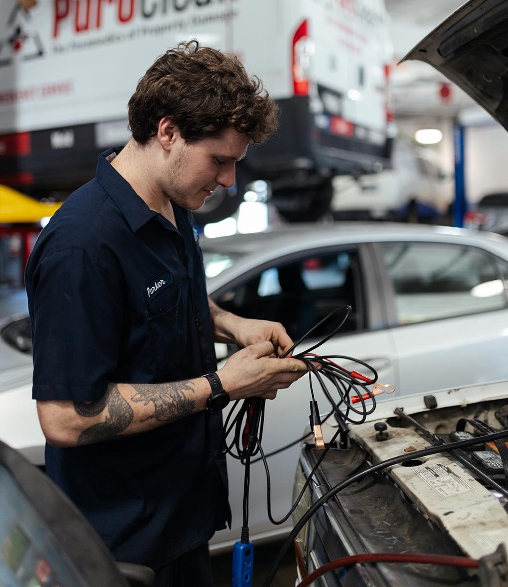 A mechanic with curly hair and tattoos on their arm, wearing a dark uniform, is working with jumper cables on a car battery at an auto repair shop.