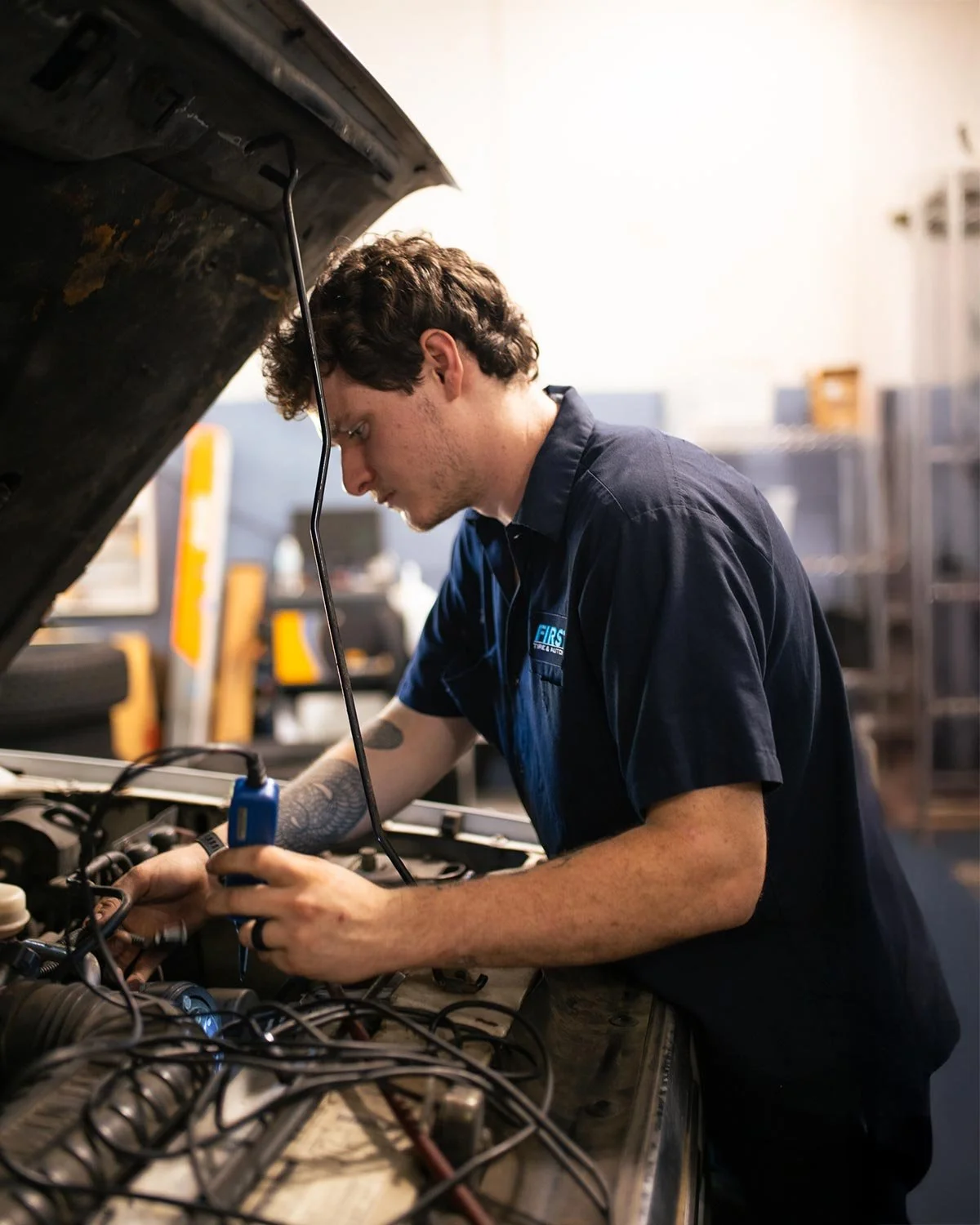 A mechanic working under the open hood of a car, using a diagnostic tool or scanner connected to the vehicle's engine, in a garage or auto repair shop.