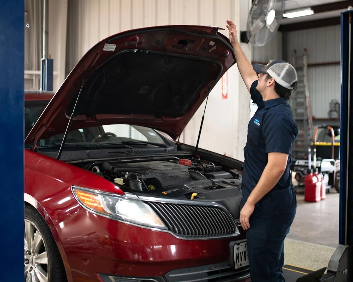 A mechanic inspecting the engine of a red car with the hood open in an auto repair shop.