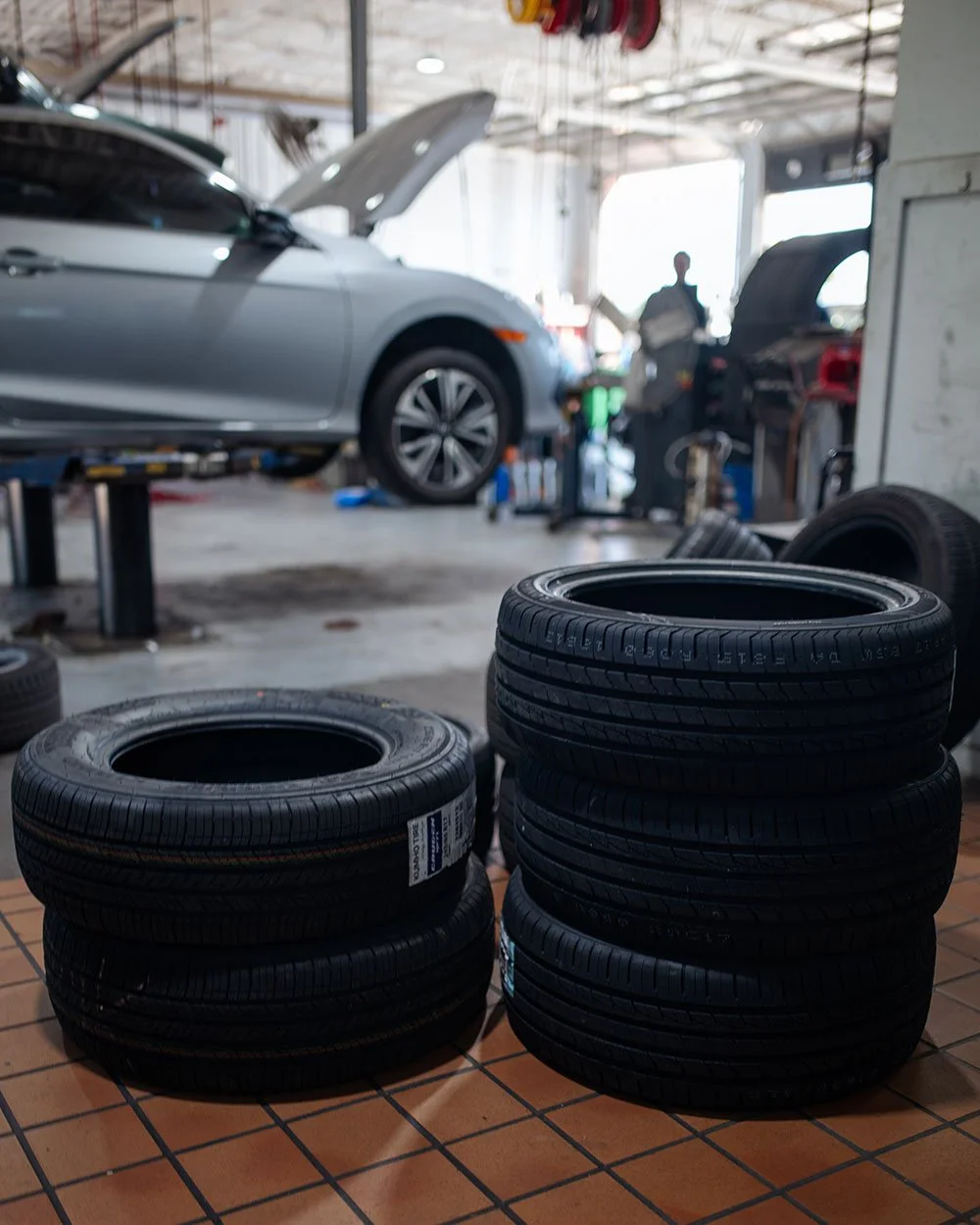 Two stacks of new tires on a tiled floor inside a busy auto repair shop, with a mechanic working on a car lifted on a lift in the background.