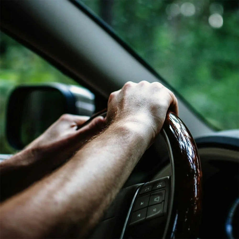 A person's hand on the steering wheel of a vehicle, driving through a green, wooded area.