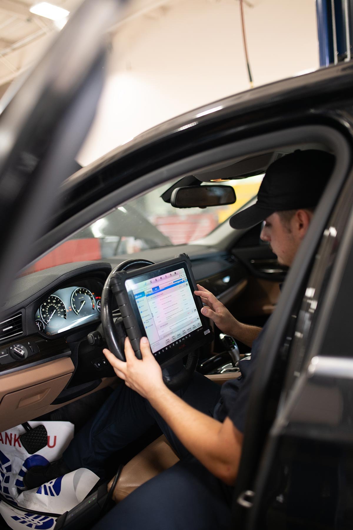A person wearing a black cap and black shirt is sitting inside a vehicle, looking at a tablet with a digital interface, while the vehicle's dashboard is visible.