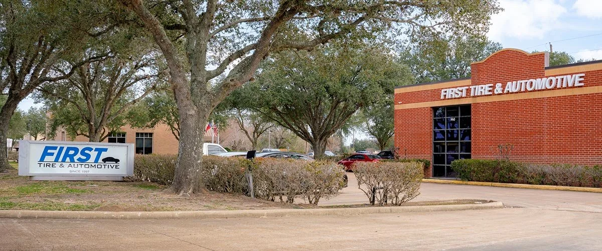 Exterior of an automotive business with a sign reading 'First Tire & Automotive,' a brick building with similar signage, trees, bushes, and parked cars.