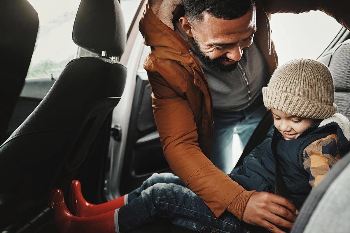 A father helping his young son into a car, with the boy sitting in a car seat, smiling and wearing a tan beanie and a dark vest.