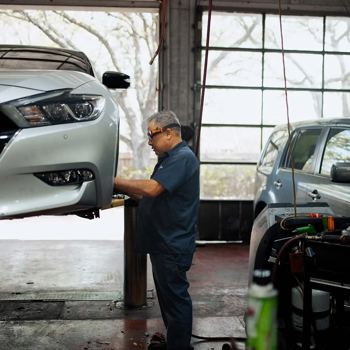 An auto mechanic working on a silver car elevated on a lift inside a garage. The mechanic is wearing safety glasses and working at a workbench. There is another vehicle in the background and equipment around.