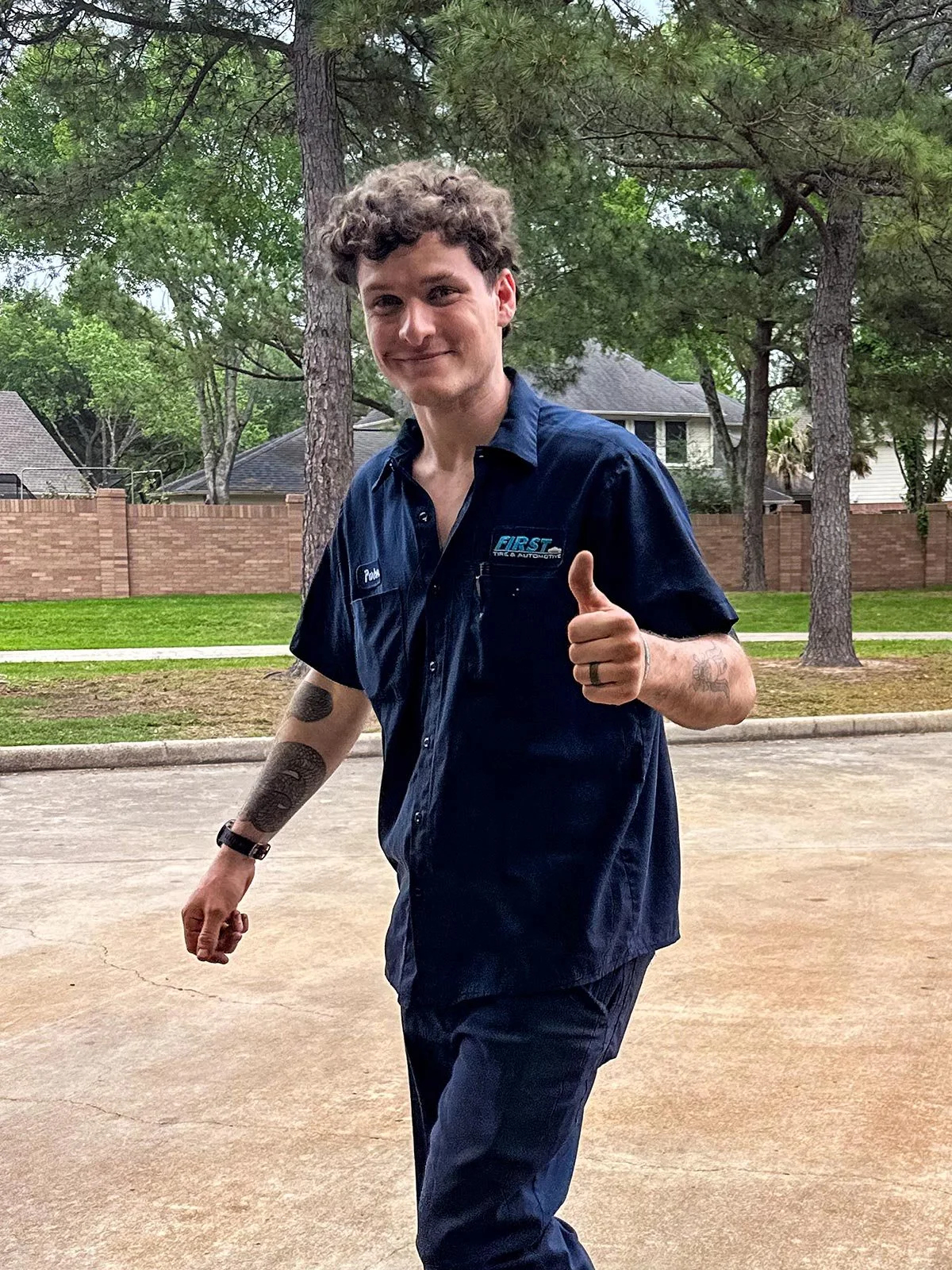 Young man with curly hair, tattoos, and a watch smiling and giving a thumbs up outdoors on a driveway, wearing a dark blue mechanic shirt with a logo and name tag.