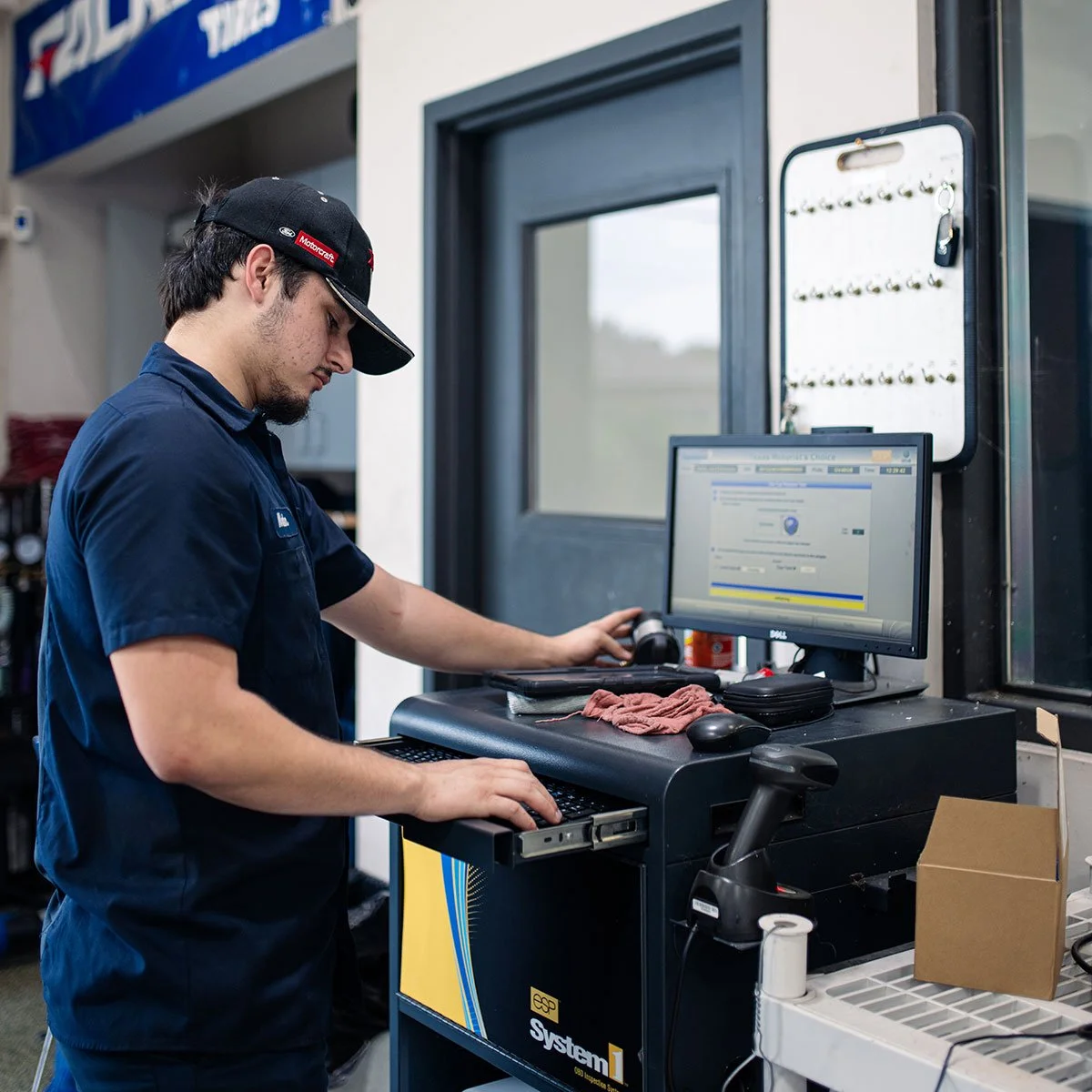 A mechanic in a blue uniform and black cap works at a computer station in an auto repair shop, with a computer monitor displaying a webpage.