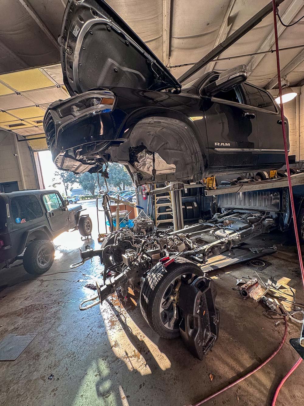 A black RAM pickup truck is lifted in an auto repair shop, with its hood open and the front end removed, exposing the engine and internal components. The vehicle's wheels are detached, and the car's body is supported by the lift.