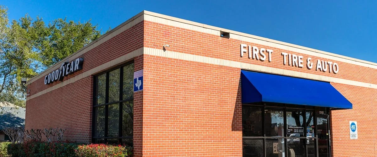 A brick commercial building with signs for Goodyear and First Tire & Auto, blue awning over entrance, large windows, and trees in the background.