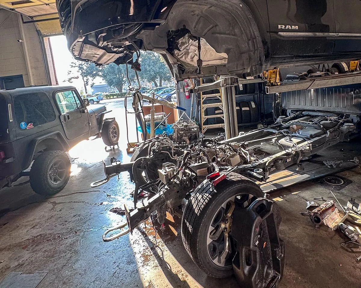 An automotive repair shop with a black RAM truck elevated, revealing its undercarriage and engine components. A disassembled vehicle frame with exposed mechanical parts is in the foreground, and a black off-road vehicle is parked nearby.