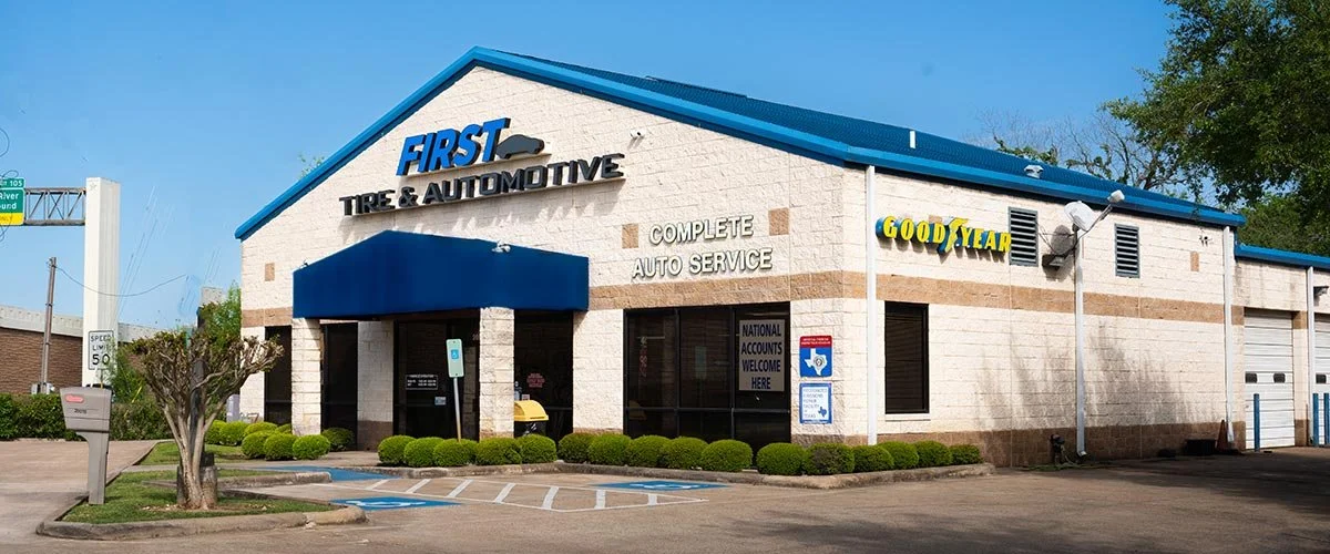 Exterior view of a tire and automotive service shop with a sign reading 'First Tire & Automotive,' a blue awning, and a blue roof. There are handicap parking spaces in front, a tree, and a few bushes. The building has a sign for 'Complete Auto Service' and 'Goodyear' branding.
