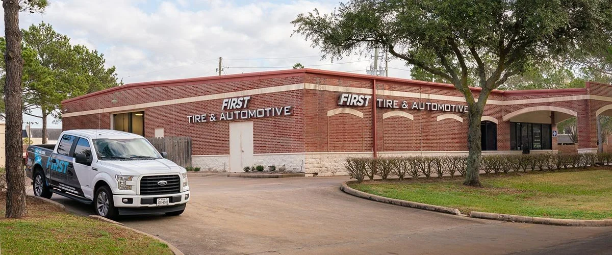 A brick building with a sign that reads "First Tire & Automotive". A white pickup truck is parked in front of the building, surrounded by green grass, trees, and a cloudy sky.