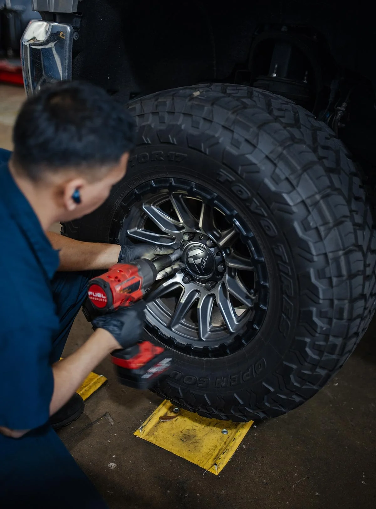 A mechanic working on a large off-road tire on a vehicle, using a power tool in a garage or workshop.