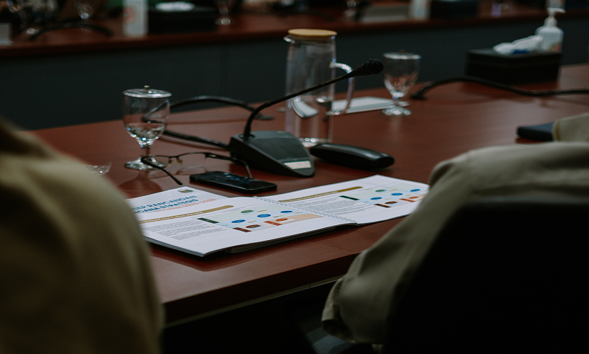 A conference room table with documents, microphones, smartphones, glasses of water, and a glass pitcher.