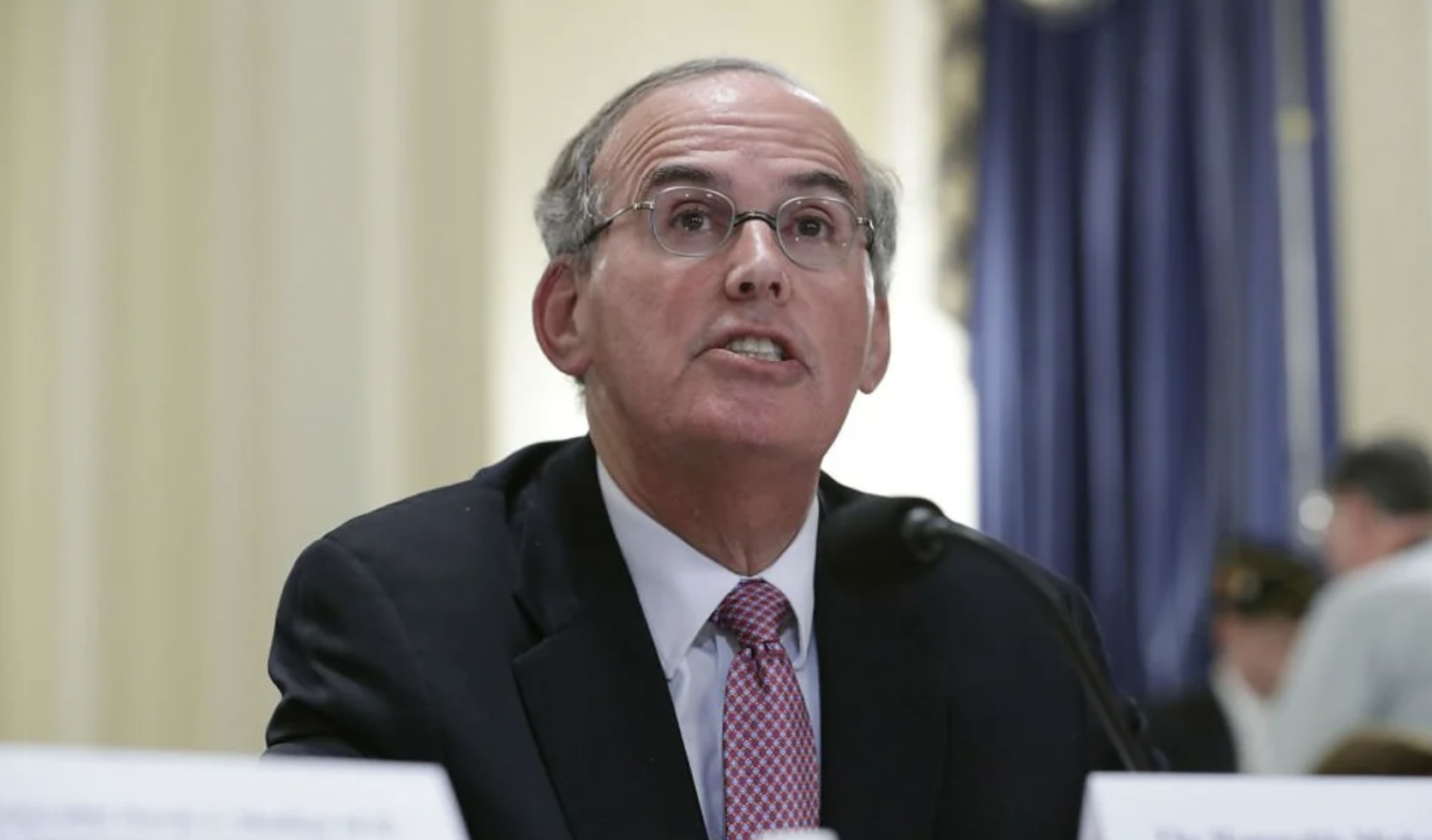 A man with glasses, gray hair, wearing a dark suit and a patterned pink tie, testifying in front of Congress with yellow walls, white columns, and blue curtains. Michael Missal, former Inspector General of Veteran's Affairs, K and L Gates lawyer.