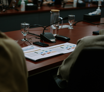 Conference table with documents, water glasses, microphones, and people seated around it.