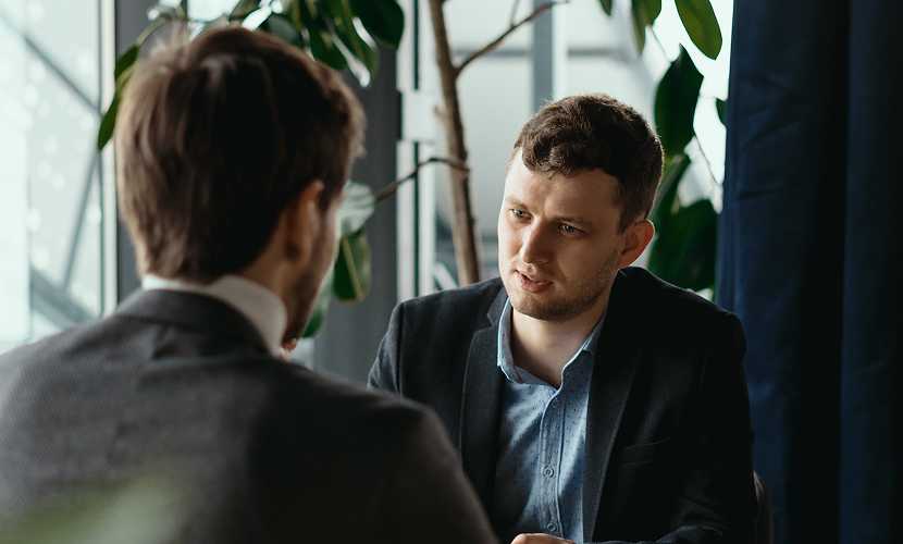 Two men engaged in a serious conversation in a modern office or conference room with large windows and green plants in the background.