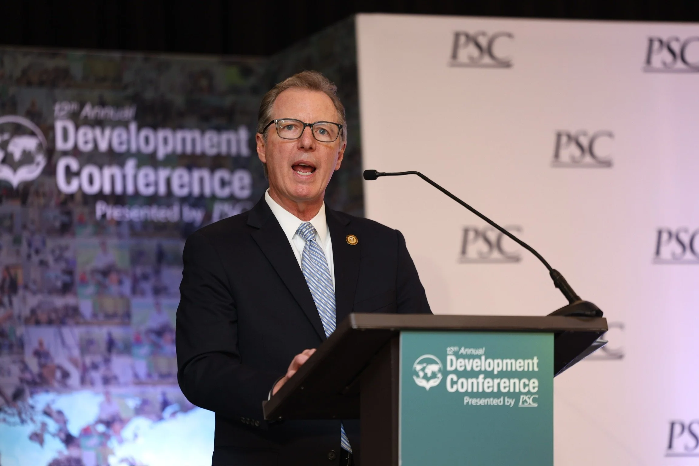 A man, Paul Martin, with glasses and a striped tie speaking at a podium during a development conference, with a backdrop displaying the conference logo and a poster with the conference title. Former Inspector General of NASA and USAID